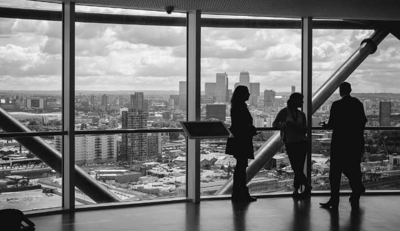 A black and white photo of people conversing on a high floor in a skyscraper with a view of the city below
